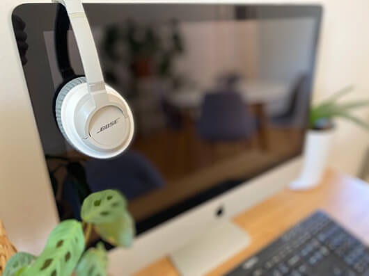 Over de rand van een zwart met zilverkleurige iMac hangt een wit met grijze Bose-koptelefoon